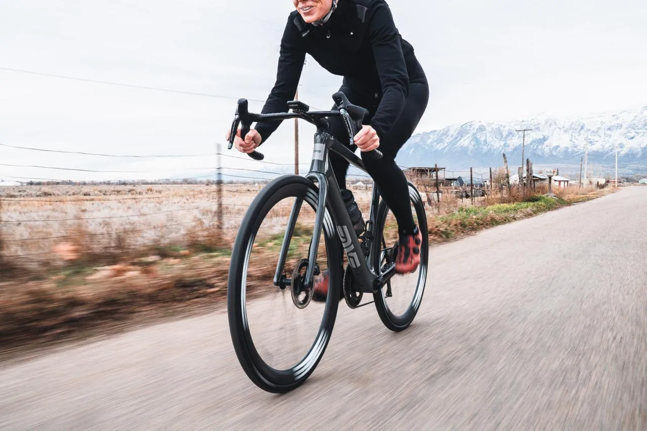 Rider on a black gravel bike with ENVE wheels sprinting along a road below snowy mountains